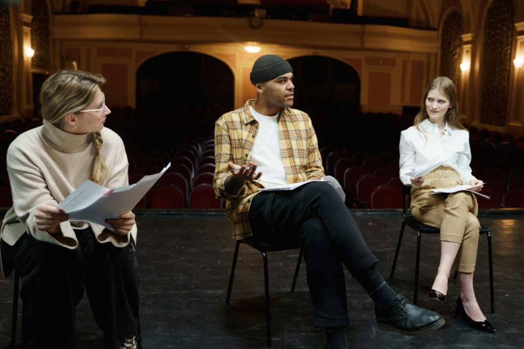 Three actors rehearsing on a theater stage, discussing scripts and performances to learn how to become an actor with no experience.