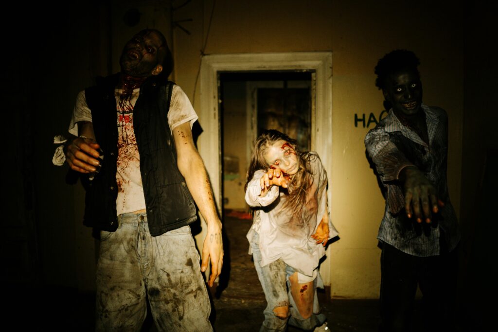 Three actors as costumed zombies outstretch their hands in a dimly lit, abandoned house setting. They are working at a haunted house scare as one of their side hustles.