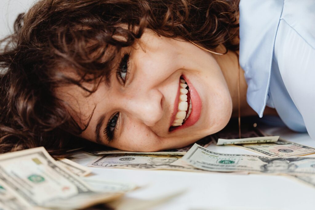 A joyful actor lies on a pile of dollar bills, smiling brightly as she invest in her acting classes and acting career.