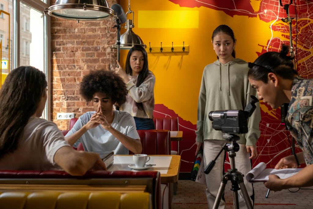 Film crew capturing a scene in a lively cafe setting with diverse actors and a cameraman for their acting classes.