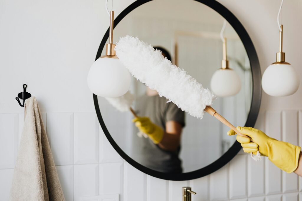 An actor at one of their side hustles cleaning an elegant bathroom mirror with a fluffy duster and yellow gloves.