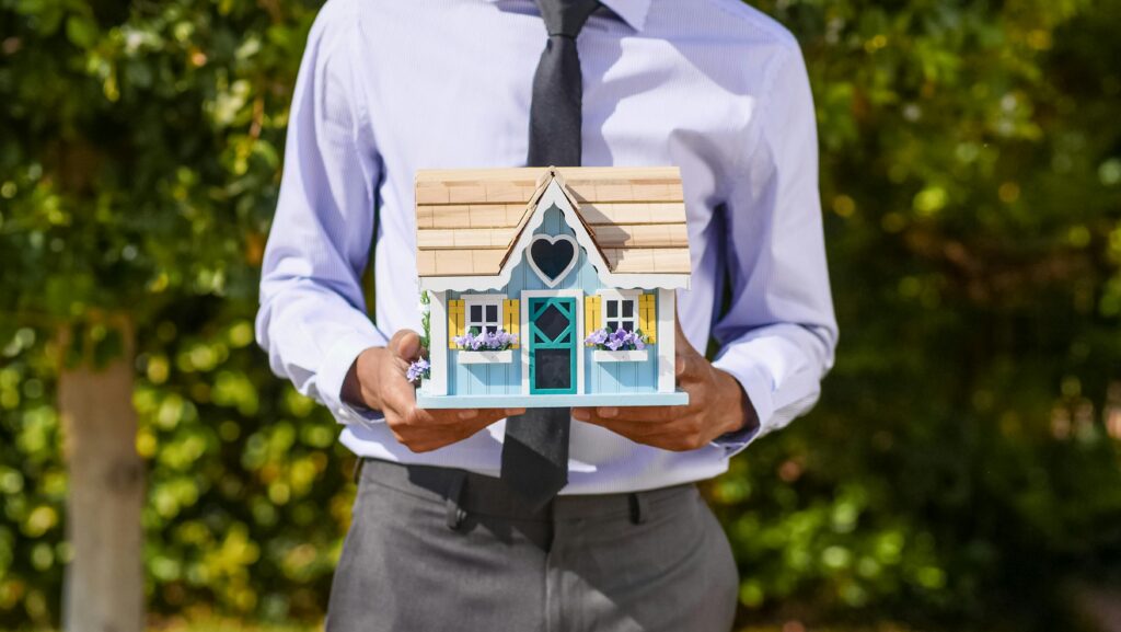 An actor and business professional holds a decorative miniature house, symbolizing real estate investment and sales as another one of their sife hustles.