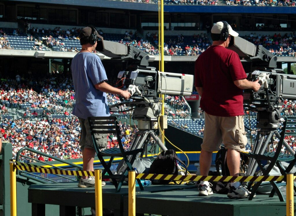 Actors working as camera operators capturing a live sports event at a crowded outdoor stadium filled with spectators to make money.