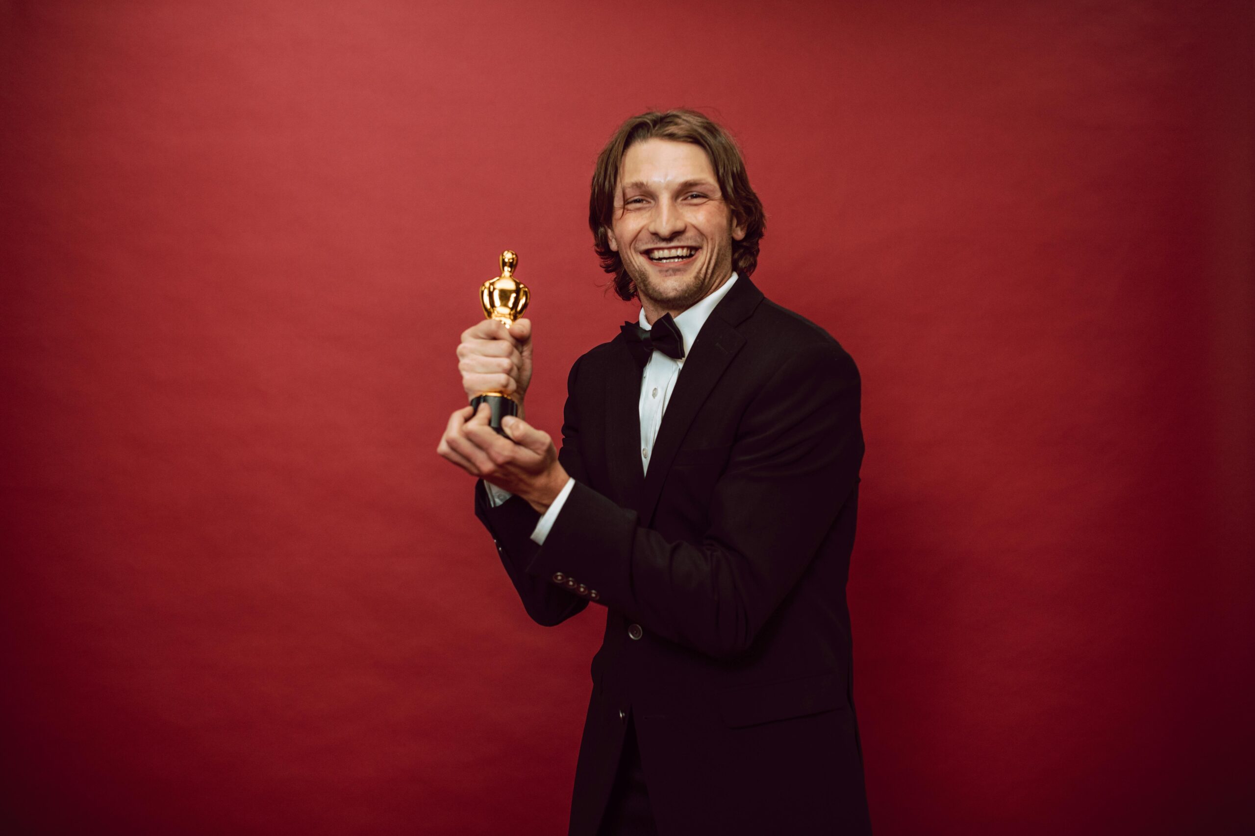 pexels-photo-7005622-7005622 Smiling actor in a tuxedo holding a trophy against a red background, celebrating success.